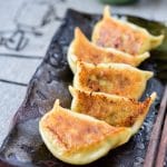 5 pan-fried shrimp dumplings on a dark stone plate with chopsticks on and a dipping sauce at the top