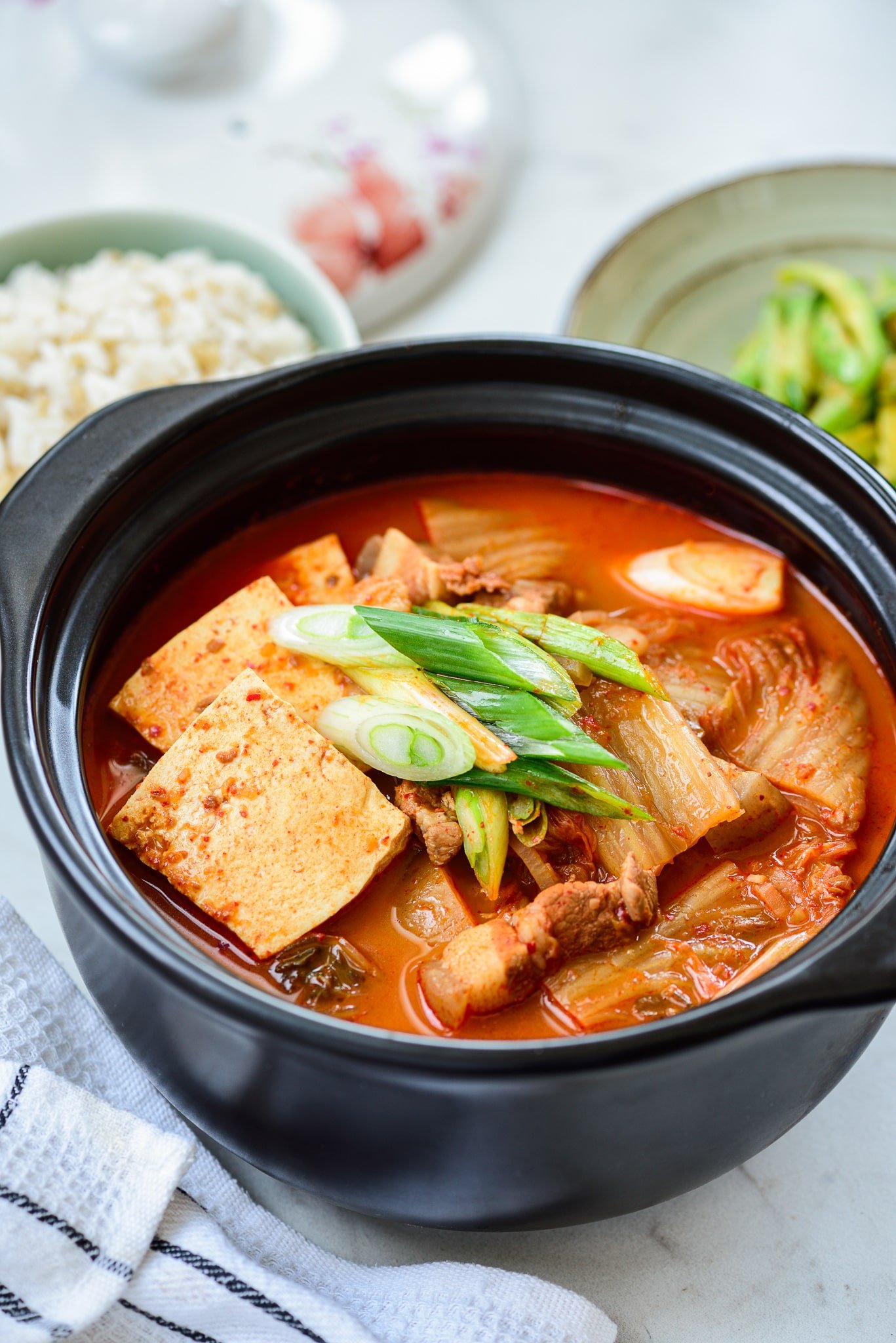 A large black pot filled with Korean kimchi jjigae stew. Bright green onions sit on top of the stew. 