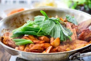 Red spicy chicken being stir-fried in a skillet with rice cakes, sweet potato, cabbage and scallions