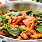 Red spicy chicken being stir-fried in a skillet with rice cakes, sweet potato, cabbage and scallions