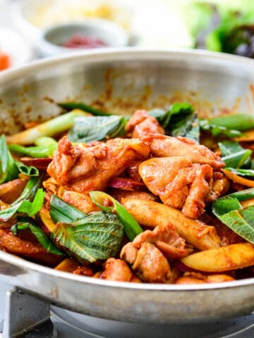 Red spicy chicken being stir-fried in a skillet with rice cakes, sweet potato, cabbage and scallions