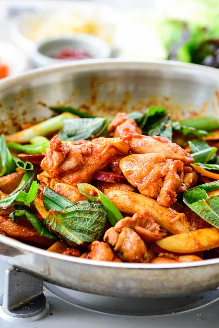 Red spicy chicken being stir-fried in a skillet with rice cakes, sweet potato, cabbage and scallions