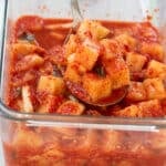 cubed radish kimchi in a glass container with a spoonful being taken out