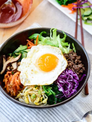 Bibimbap served in a large black bowl with various vegetables and meat toppings with a sunny side up fried egg on top and a gochujang sauce in a small glass bowl on the side