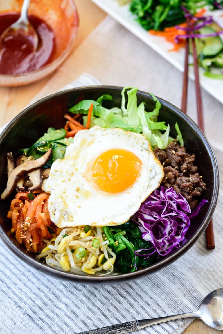 Bibimbap served in a large black bowl with various vegetables and meat toppings with a sunny side up fried egg on top and a gochujang sauce in a small glass bowl on the side