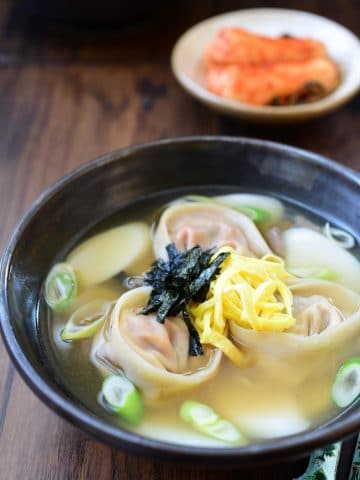 a bowl of Korean rice cake soup with kimchi dumplings garnished with egg omelette strips served in a large black bowl with a small kimchi side dish in the background